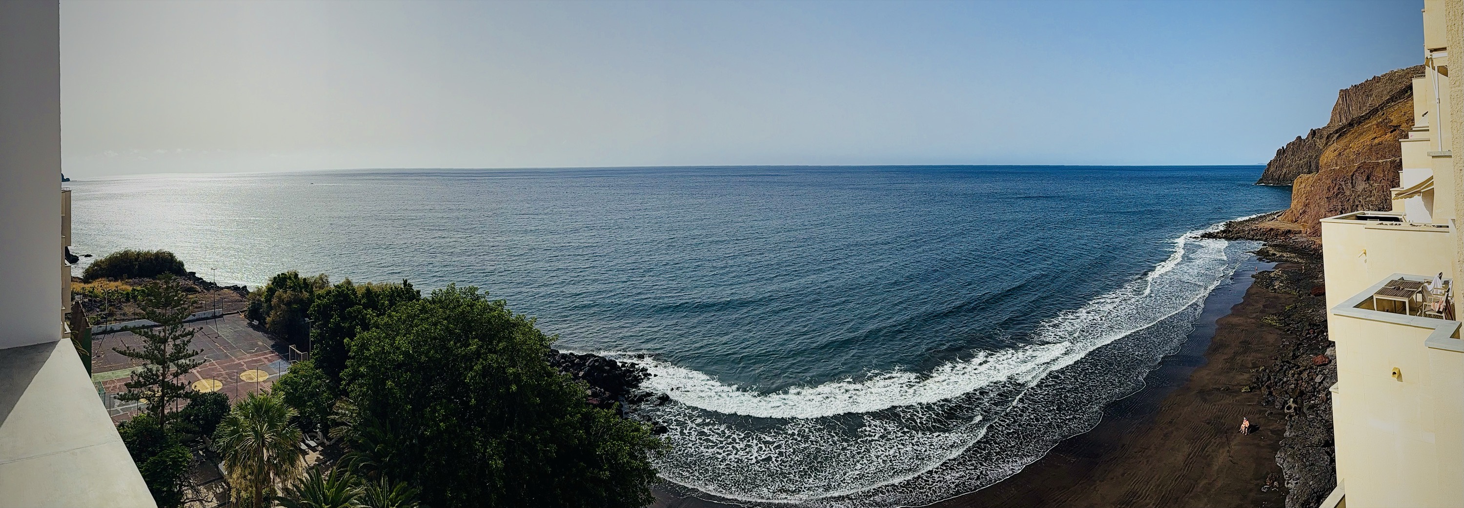 Panorama from the ninth-floor terrace — a dark-sand crescent directly below, white surf, deep Atlantic, red-ochre cliffs to the right